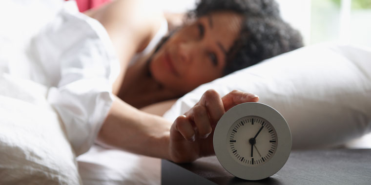 Woman lays in her bed, touching her alarm clock during daylight saving time
